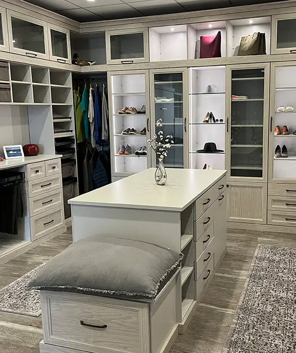 Elegant walk-in closet featuring a center island with drawers and a vase of flowers, a cushioned bench, glass-front shoe and accessory cabinets