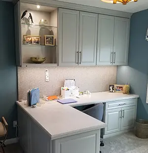 Custom-built home office featuring an L-shaped white desk with cabinetry above and below, set against a textured backsplash and soft blue walls