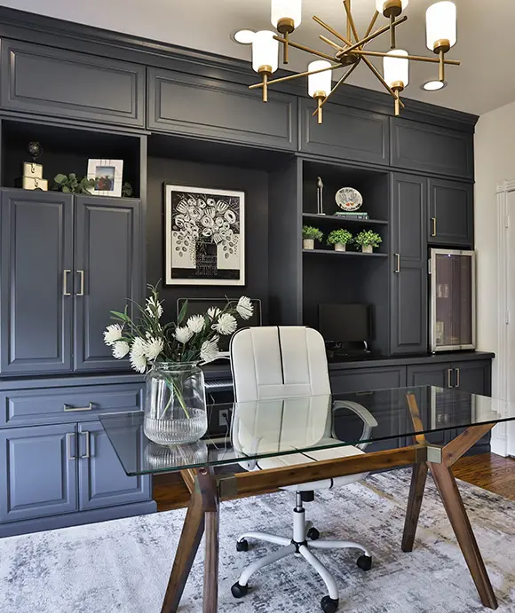 Sophisticated home office featuring floor-to-ceiling navy built-ins, a white swivel chair, and a glass desk with a wood base, accented by a modern gold chandelier and fresh white flowers in a glass vase
