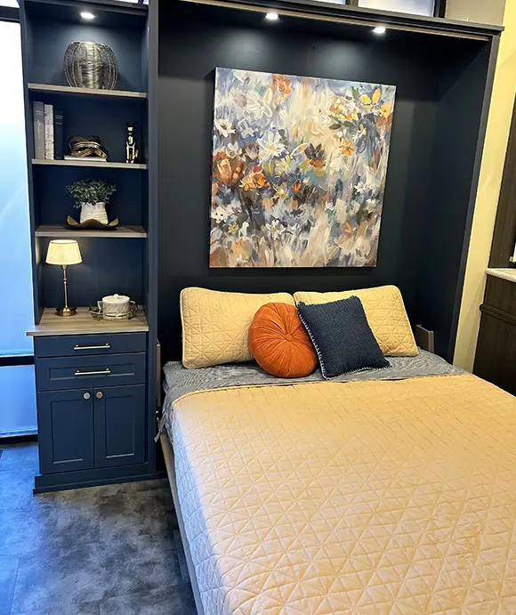 Navy blue wall bed with built-in shelving and drawers, featuring a cozy beige quilt, colorful accent pillows, and a floral painting above the headboard