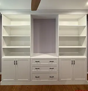 Built-in white wall unit featuring open shelving on both sides, a recessed center section, three central drawers with black handles, and lower cabinets for additional storage