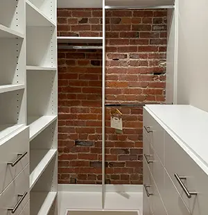 Contemporary walk-in closet with sleek white cabinetry and shelving, modern metal handles, and a striking exposed red brick accent wall