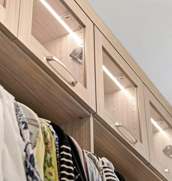 Overhead glass-front cabinets with built-in LED light strips above hanging wardrobe section in a custom closet.