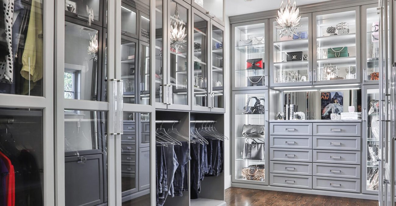 Modern glam closet in soft gray with glass-front cabinetry, integrated lighting, display shelving for handbags and jewelry, and double-hang sections with chrome rods.