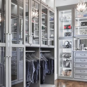 Modern glam closet in soft gray with glass-front cabinetry, integrated lighting, display shelving for handbags and jewelry, and double-hang sections with chrome rods.