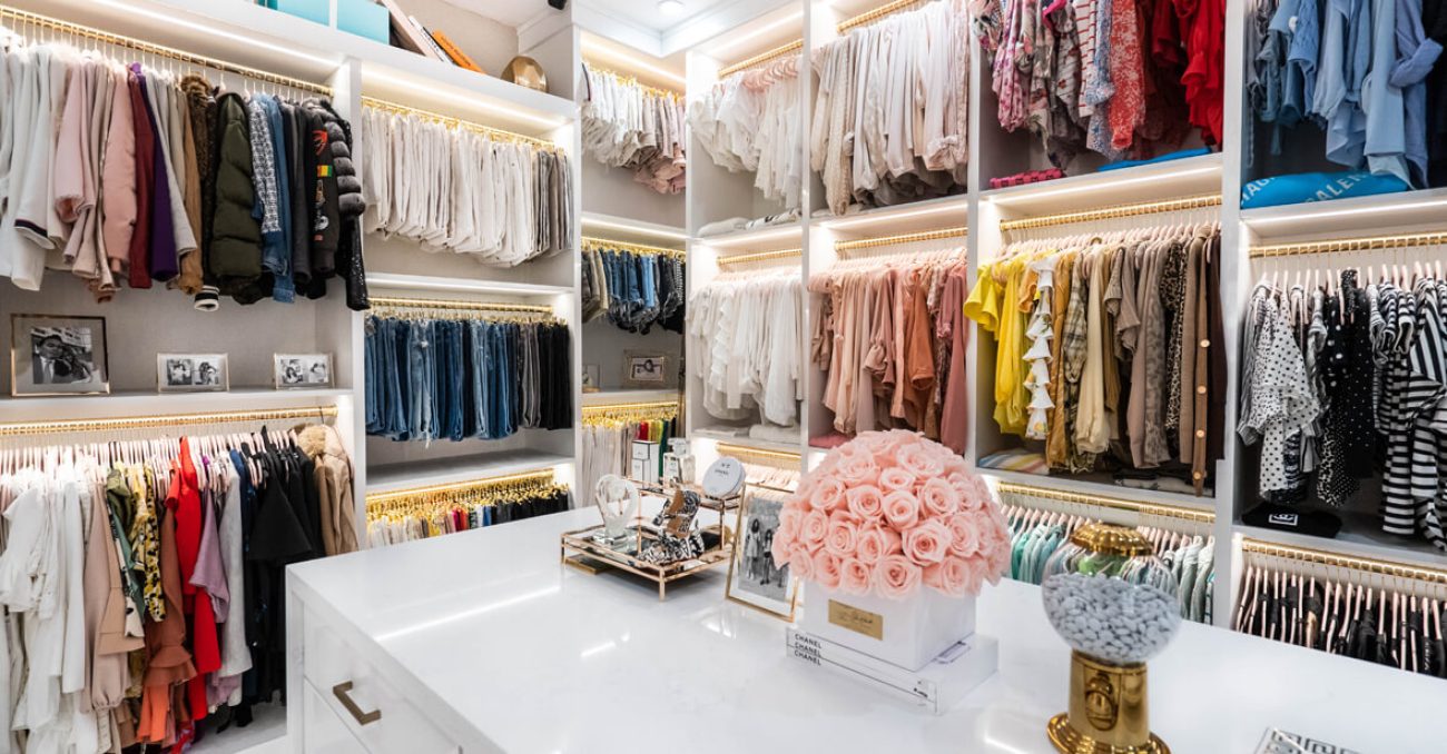 Custom white melamine system with triple-hang wardrobe rods, gold-tone hanging hardware, and integrated LED lighting. Central island provides additional accessory organization space.