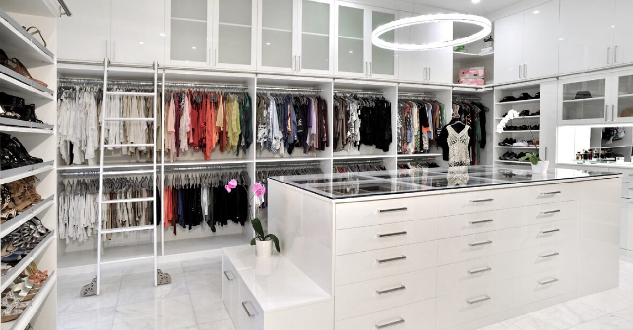 Custom closet in white high gloss, featuring full-height hanging sections, glass display island with built-in drawers, integrated ladder rail, and overhead cabinets with frosted glass fronts.