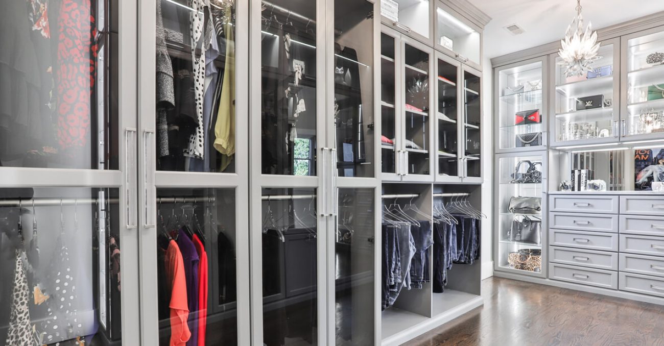 Glass-front wardrobe system in Dove Grey with integrated LED lighting, double-hang rods, and display shelving. Features stacked drawer banks, acrylic shelf dividers, and a mirrored vanity station.