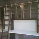 Custom home office featuring dark wood cabinetry, glass display cabinets, open shelving, a large workstation, and a rolling library ladder.
