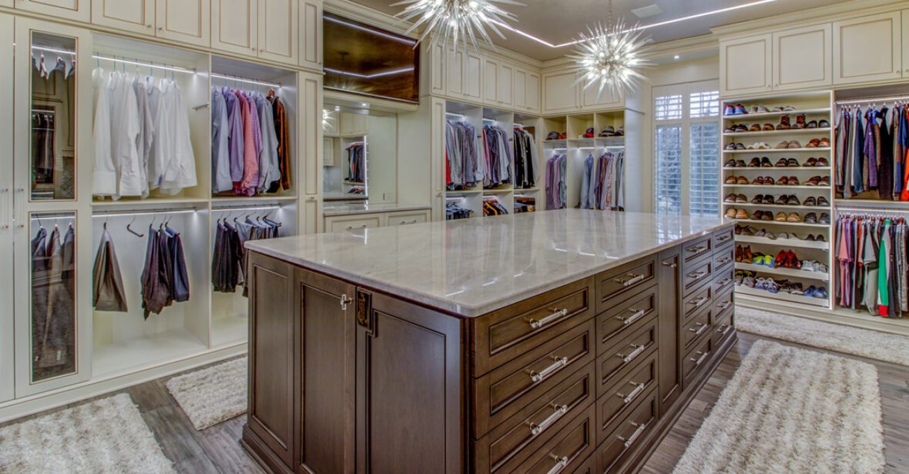 Spacious closet with cream-toned cabinets, a large island with deep drawer storage, built-in glass shoe shelving, and double hanging rods.