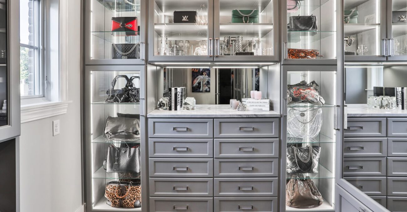 Modern gray closet with integrated lighting, glass display shelves for designer handbags, and a dedicated vanity station with full-extension drawers.