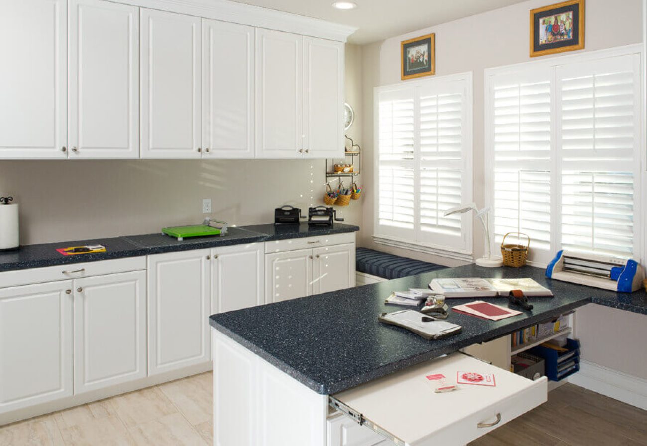 Custom craft room Raised Panel cabinet doors in Antique White melamine with a dark solid-surface countertop. Features a slide-out ironing drawer, lower open shelving, and wraparound work surface.