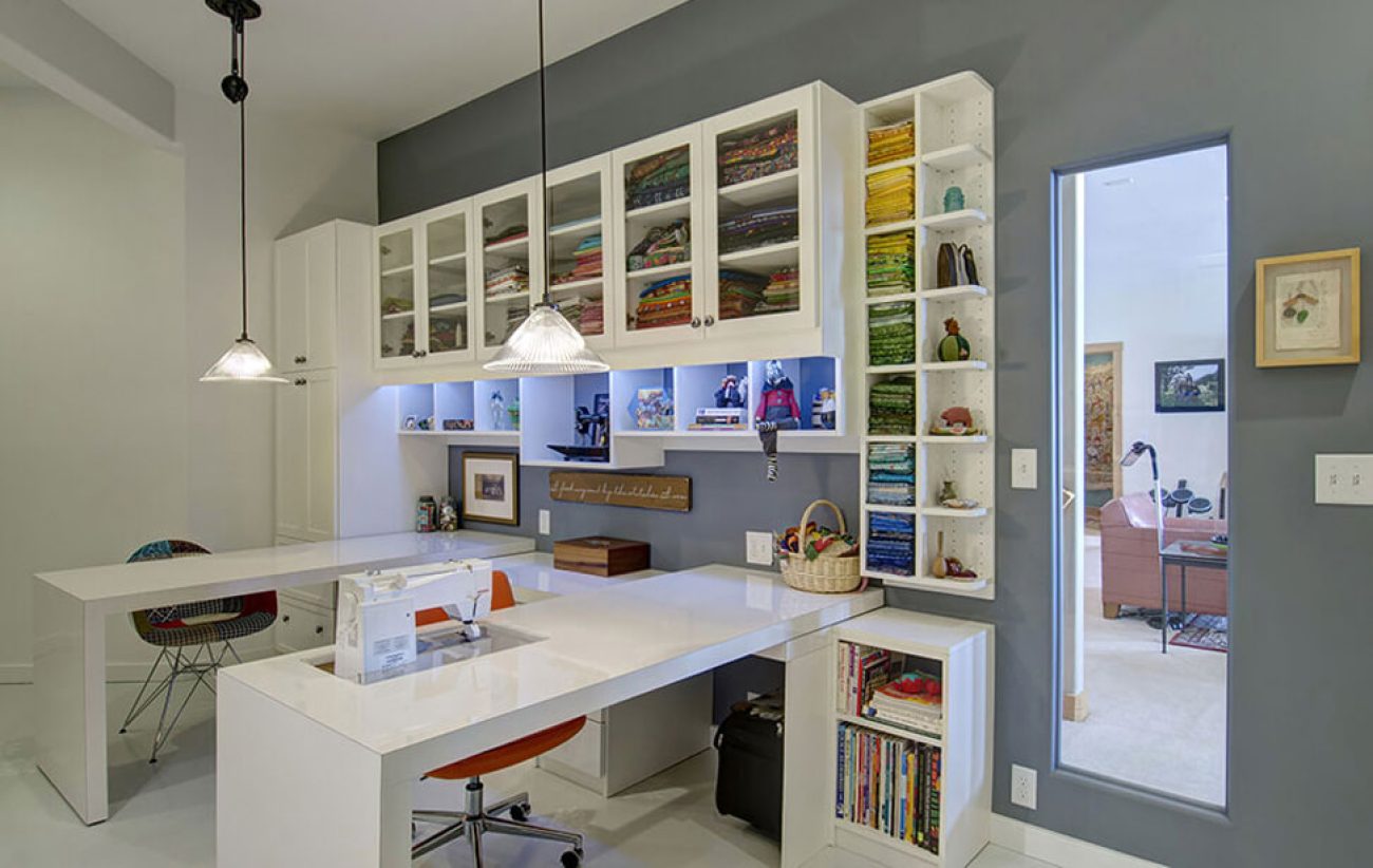 Craft room organization with Glass-front upper cabinets with Shaker frames in White Chocolate melamine. Includes vertical fabric cubbies, open book shelving, and a built-in sewing workstation with wraparound desk.