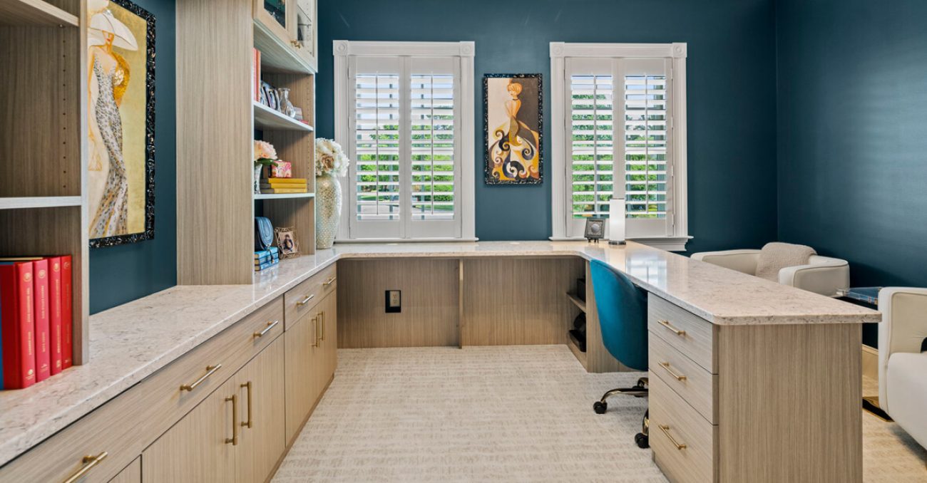 Light wood-finish home office cabinetry surrounds an L-shaped quartz desk, featuring built-in bookshelves