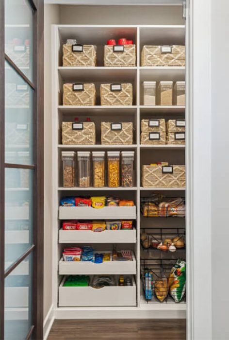 Narrow walk-in pantry optimized with labeled woven bins, cereal containers, snack drawers, and pull-out wire baskets for efficient small space storage.