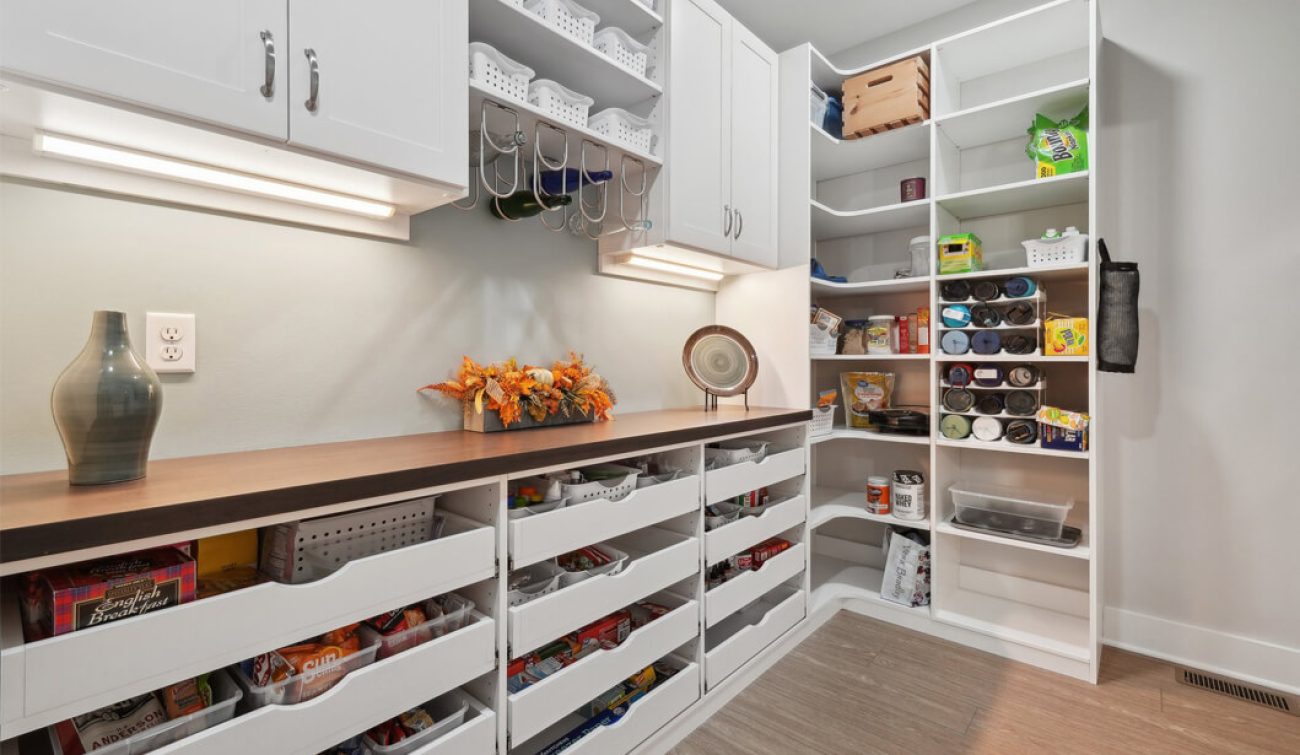 Custom white pantry with multiple open pull-out drawers and upper cabinetry, featuring a wood slab countertop and wine glass holders.
