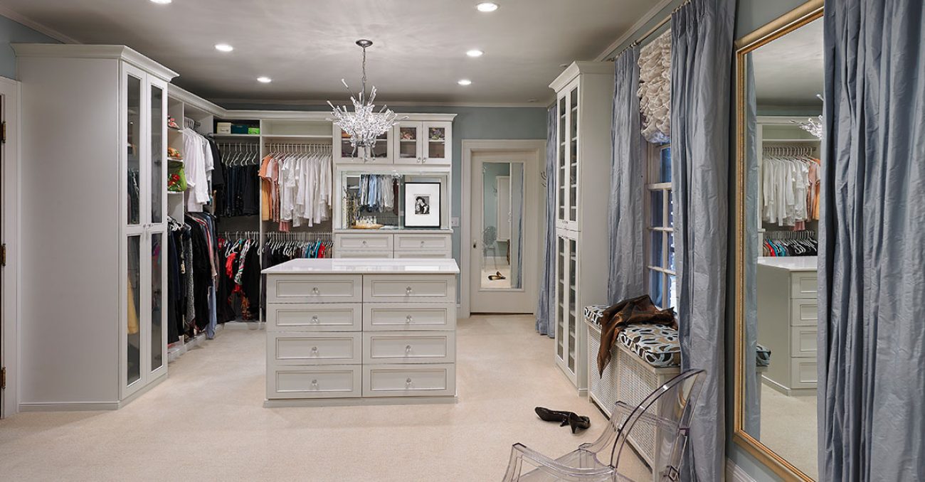 Expansive dressing room featuring a central island with drawer storage, dual-level hanging rods, integrated wardrobe cabinetry with glass doors, and upper storage